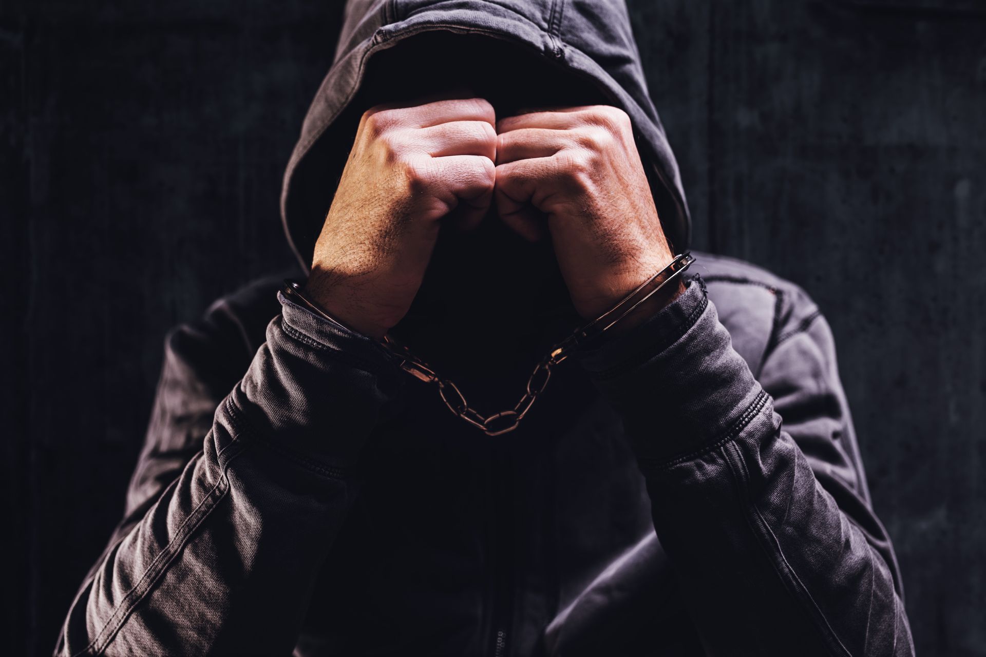 Hooded person with hands clasped over face in a dark, moody setting