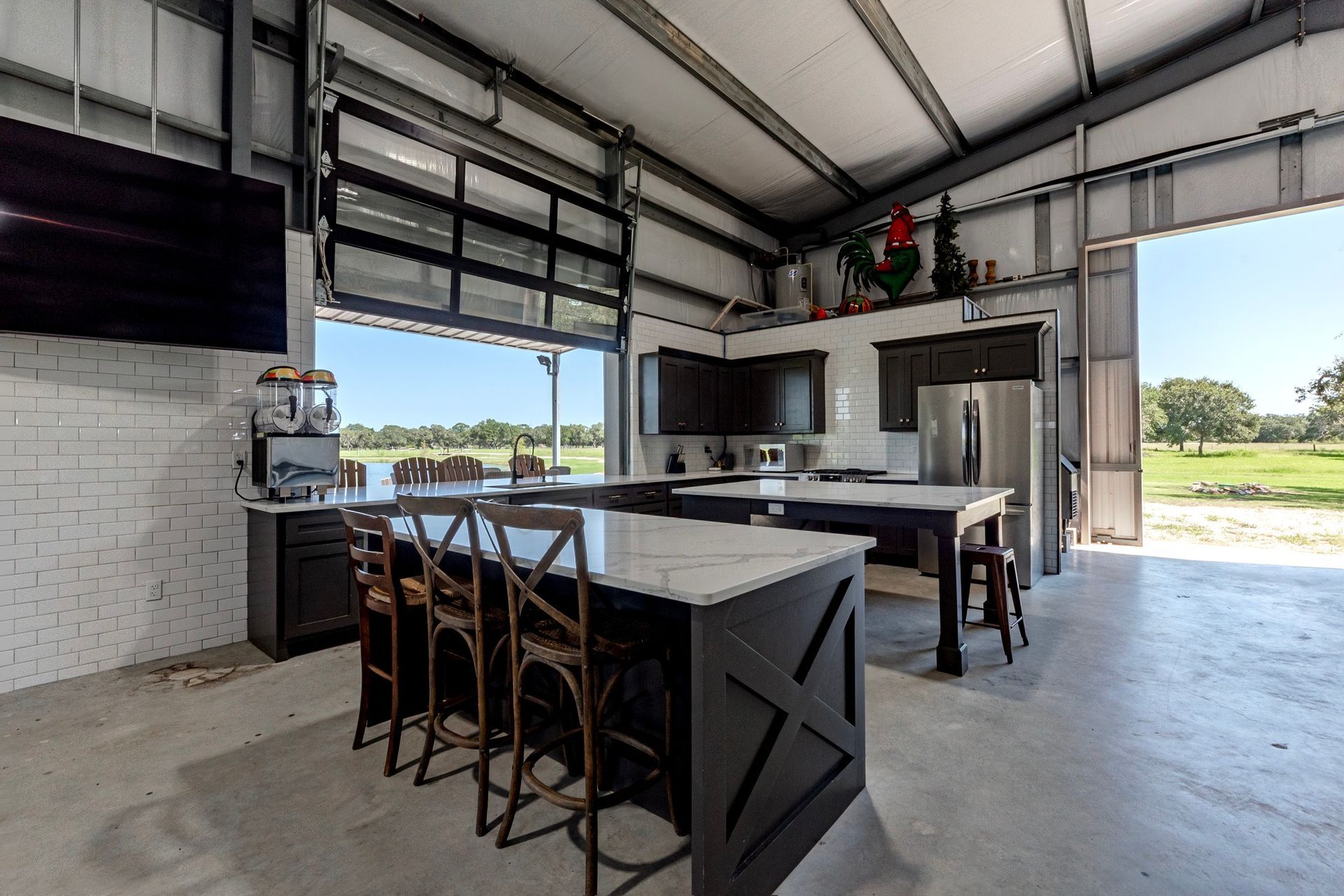 Kitchen in a steel-frame building, with an open garage door view, large island, stainless steel appliances, dark cabinets, and outdoor view.
