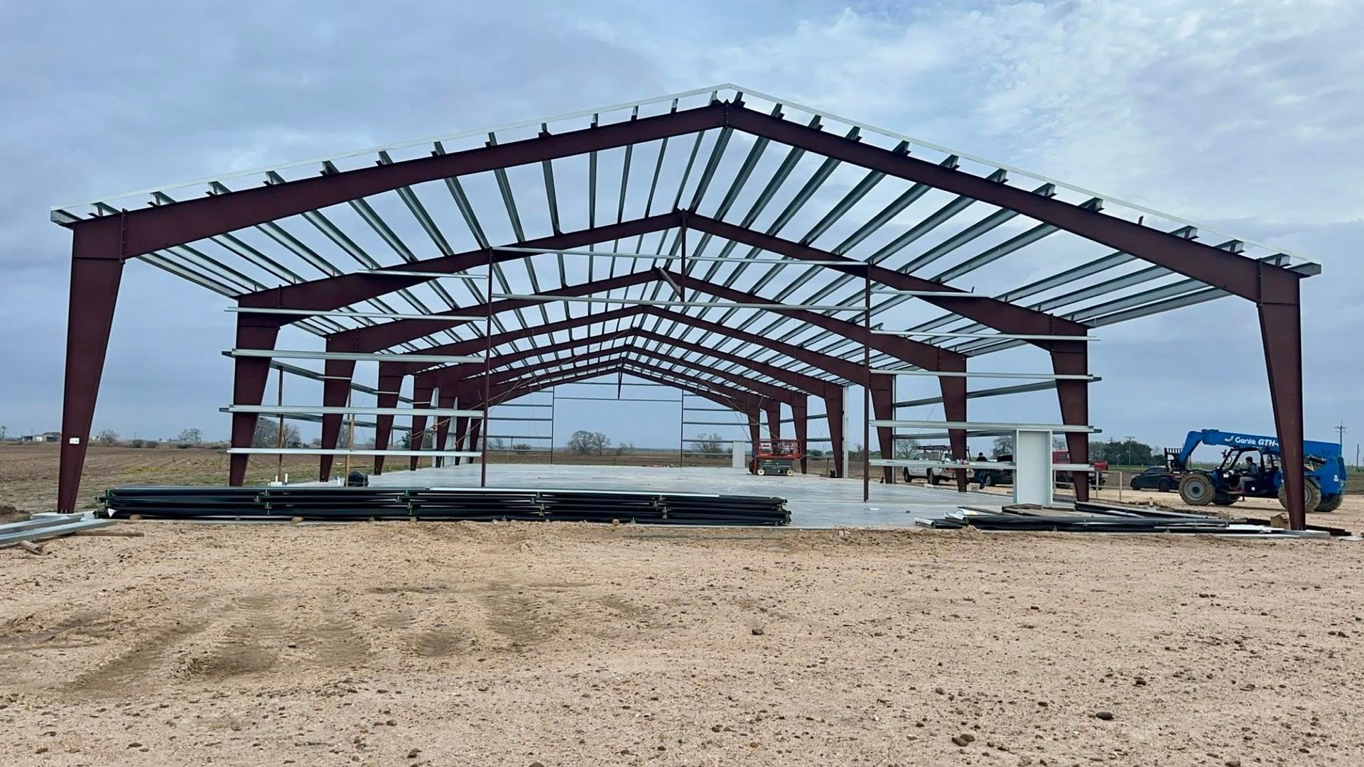 Metal frame of a building under construction, brown with a grey concrete base, in an open field.