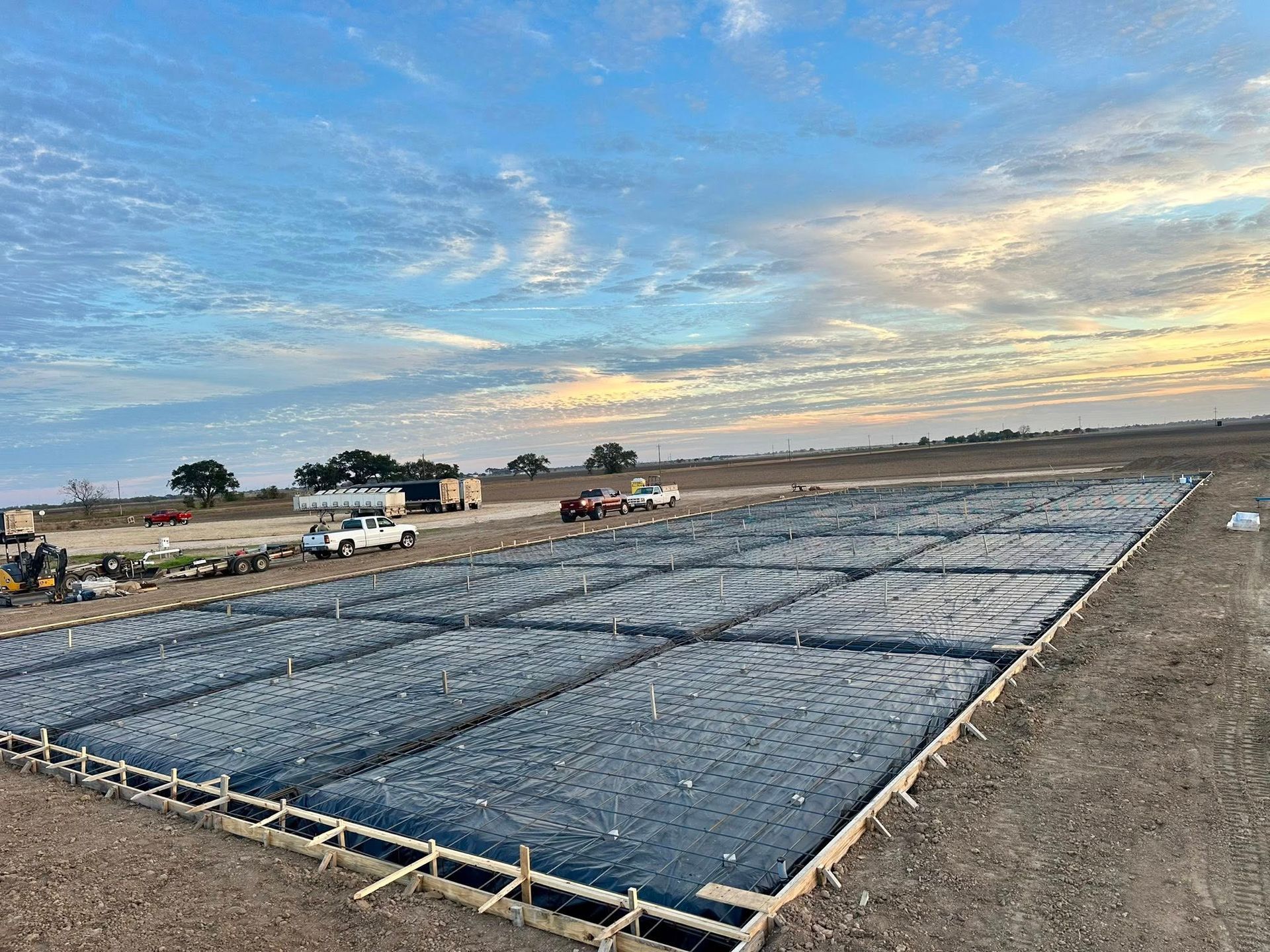 Construction site with a large concrete form, covered in black sheeting, against a field under a cloudy sky.