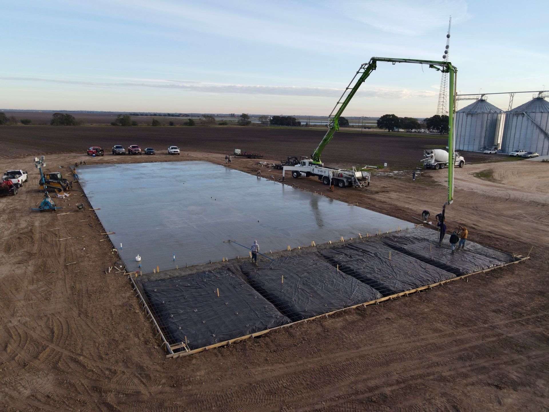 Construction site: Concrete being poured for a large rectangular foundation, using a pump truck. Rural setting.