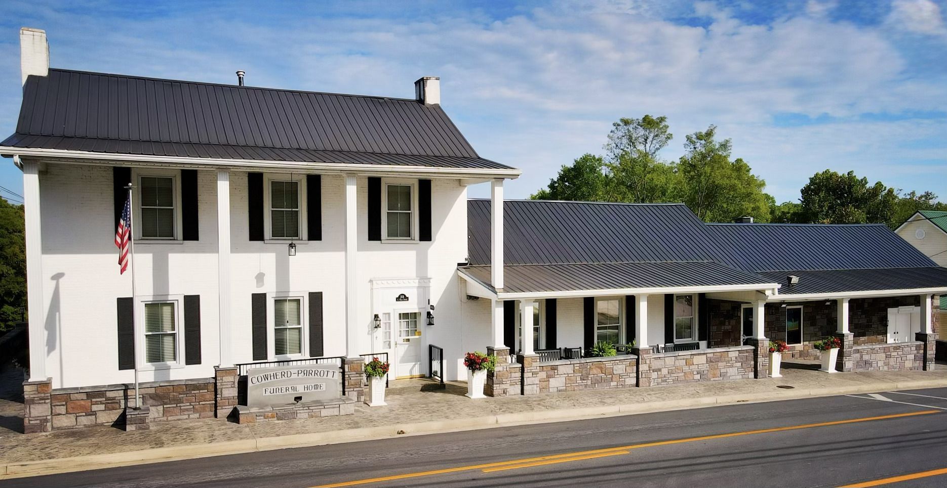 White two-story building with a covered porch and American flag by a roadside under a blue sky