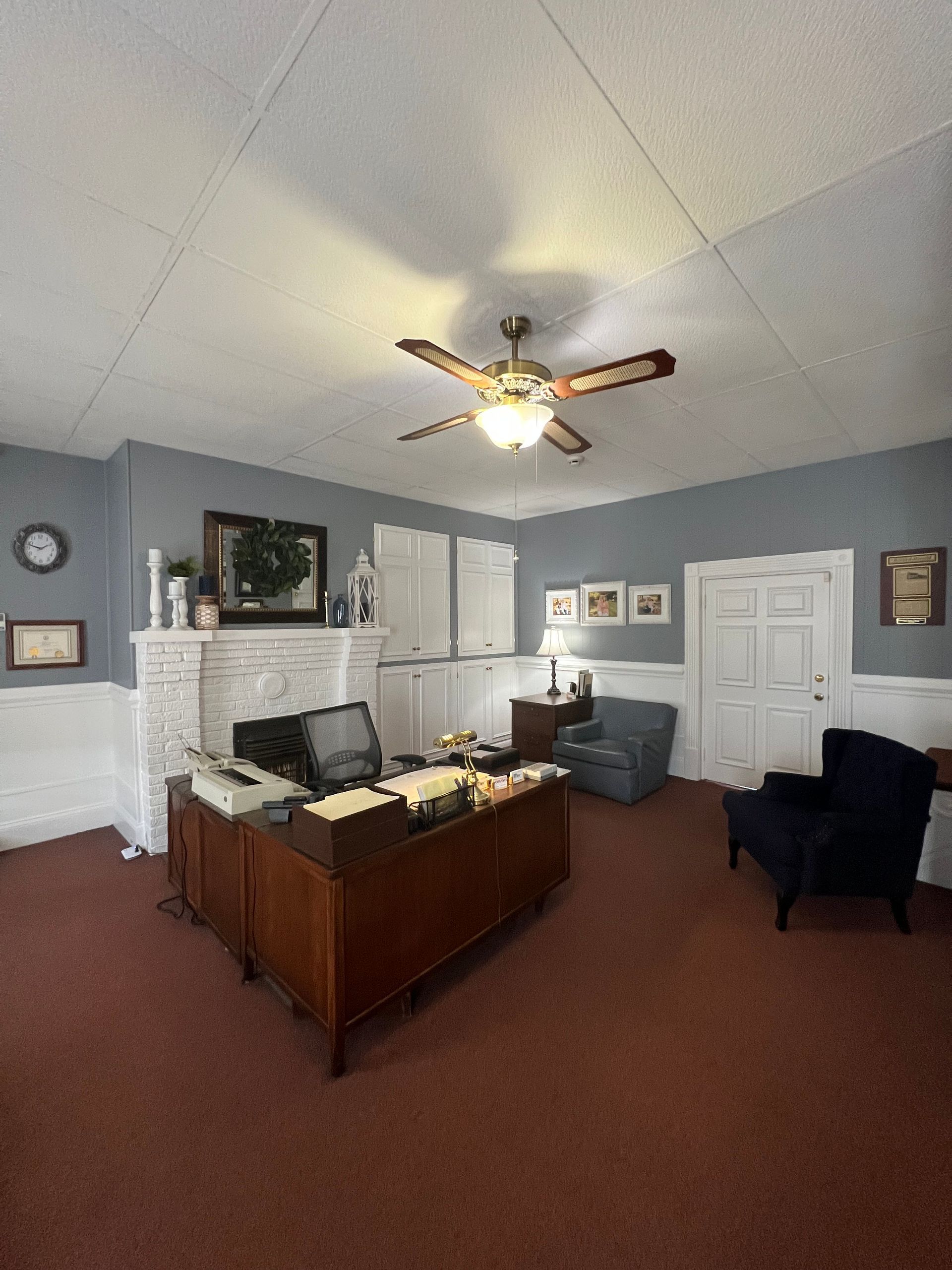 Office with white fireplace, wooden desk, gray walls, red carpet, and ceiling fan under a vaulted ceiling