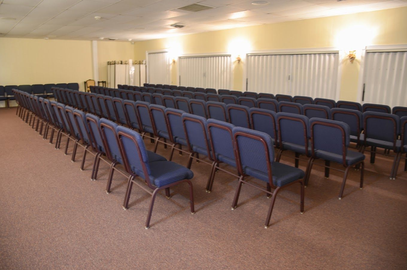 Empty conference room with rows of blue chairs facing the front, beige walls, and windows with blinds.