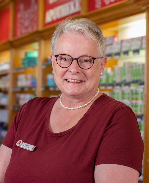 Eine Person mit kurzen, hellen Haaren und Brille, die ein weinrotes Hemd und eine Perlenkette trägt, steht in einer Apotheke.