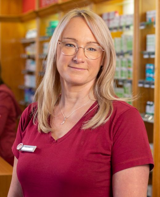 Eine Person in einem bordeauxroten Kasack mit V-Ausschnitt und Brille steht in einer Apotheke; im Hintergrund sind Regale mit Produkten zu sehen.