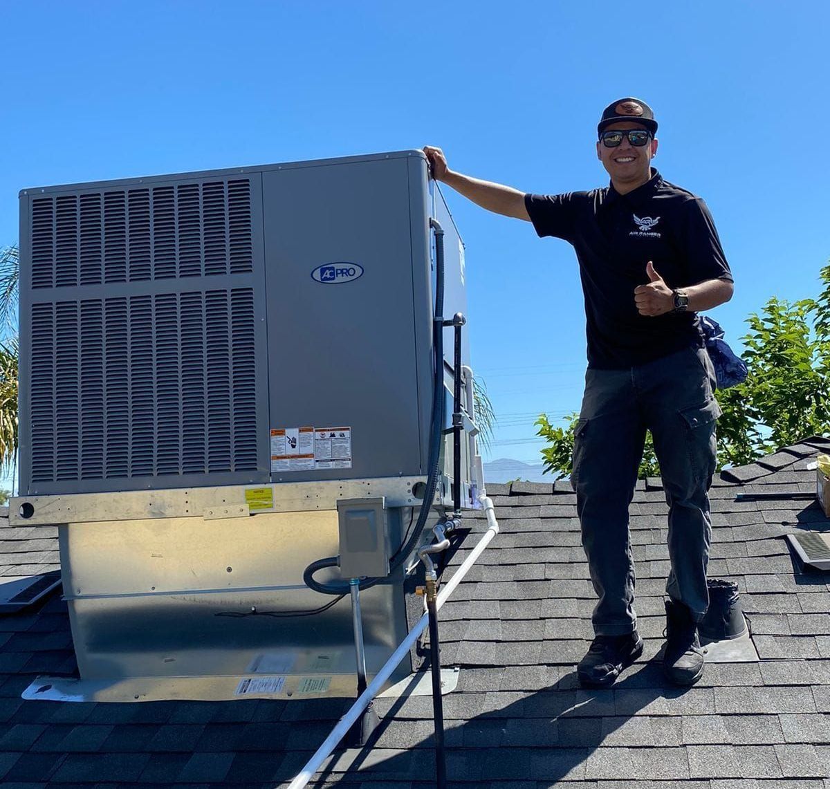 A man is standing on a roof next to an air conditioner and giving a thumbs up.