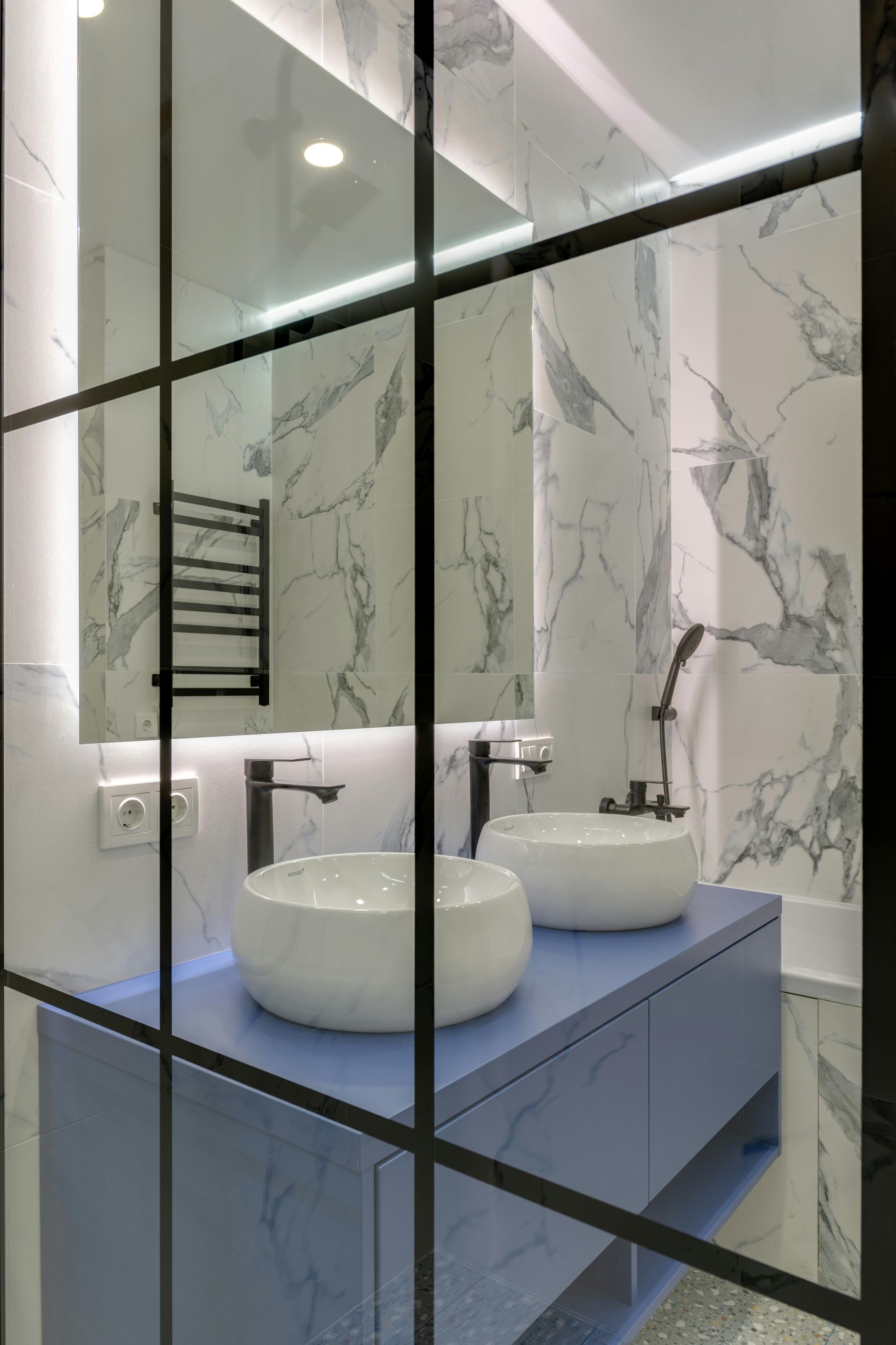 Bathroom with a blue vanity, marble walls, two white sinks, and a black-framed mirror.