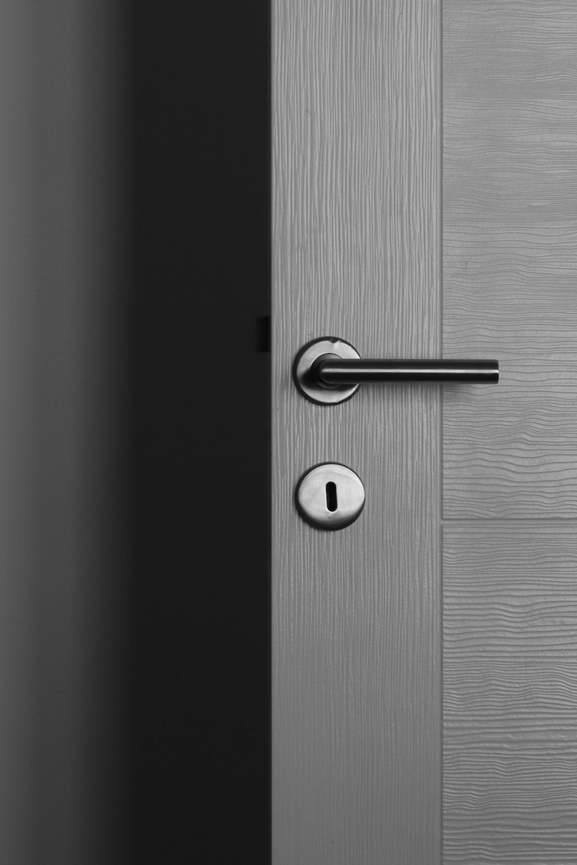 Partially open light-colored wooden door with dark handle and keyhole, revealing a dark interior.