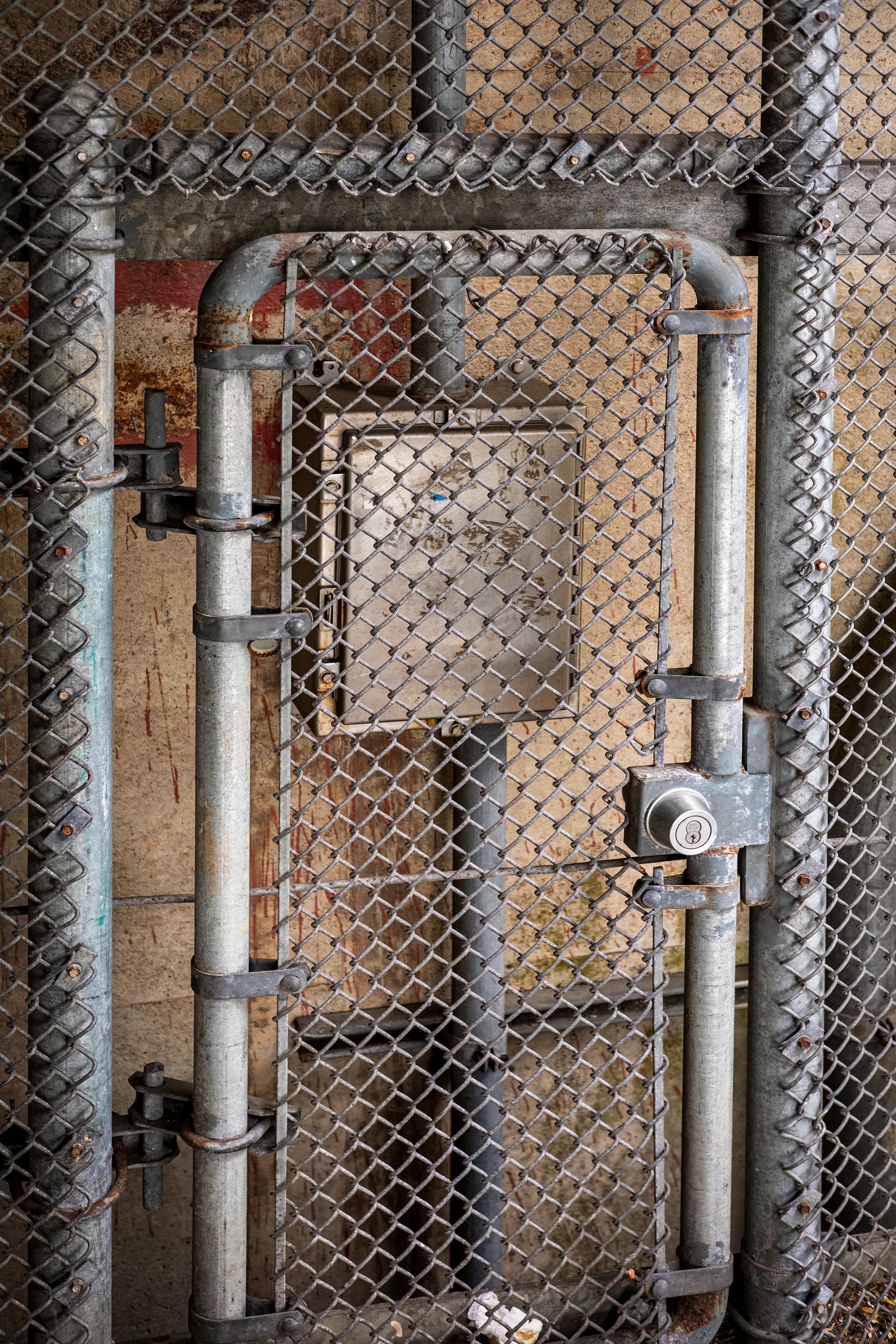 A rusty chain link gate with a lock, protecting a weathered electrical box in an industrial setting.