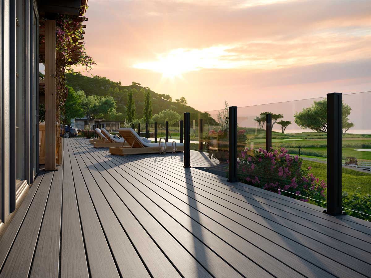 Trex® Decking from Budget Home Supply Colorado - Wooden deck with black posts and glass railing overlooking a green landscape and ocean at sunset.