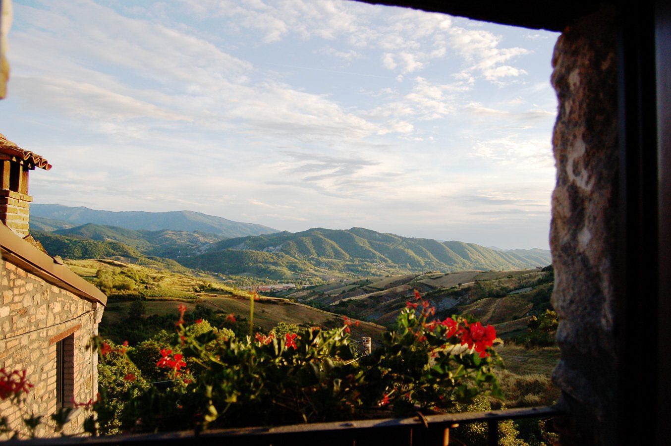 View of rolling green hills and mountains from a stone window with red flowers.