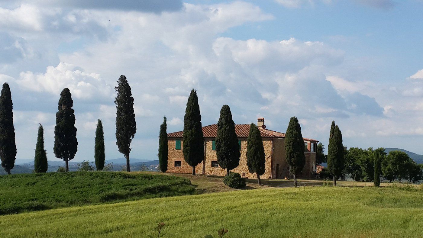 Stone farmhouse surrounded by tall cypress trees and green fields under a partly cloudy sky.