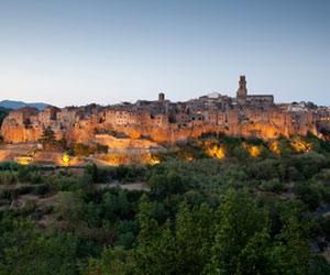 Hilltop medieval town glowing with warm lights at dusk, surrounded by dense greenery.
