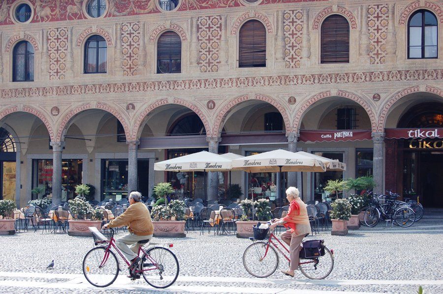 Two people ride bicycles past an ornate Italian café with arched colonnades and outdoor seating.