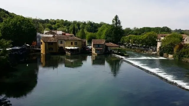 Cluster of rustic buildings on a riverbank surrounded by trees, with a small dam creating gentle ripples in the water.
