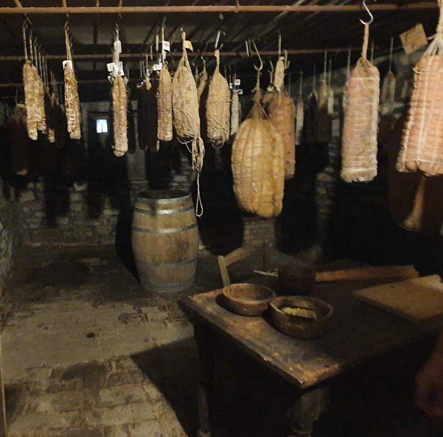 Dimly lit cellar with various cured meats hanging from the ceiling, a wooden barrel, and an old table with bowls.