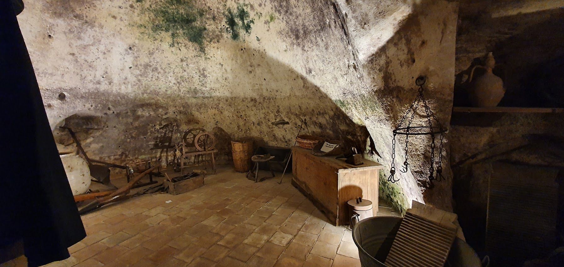 A dimly lit cave dwelling with rustic wooden furniture, antique tools, and stone walls covered in moss and patina.