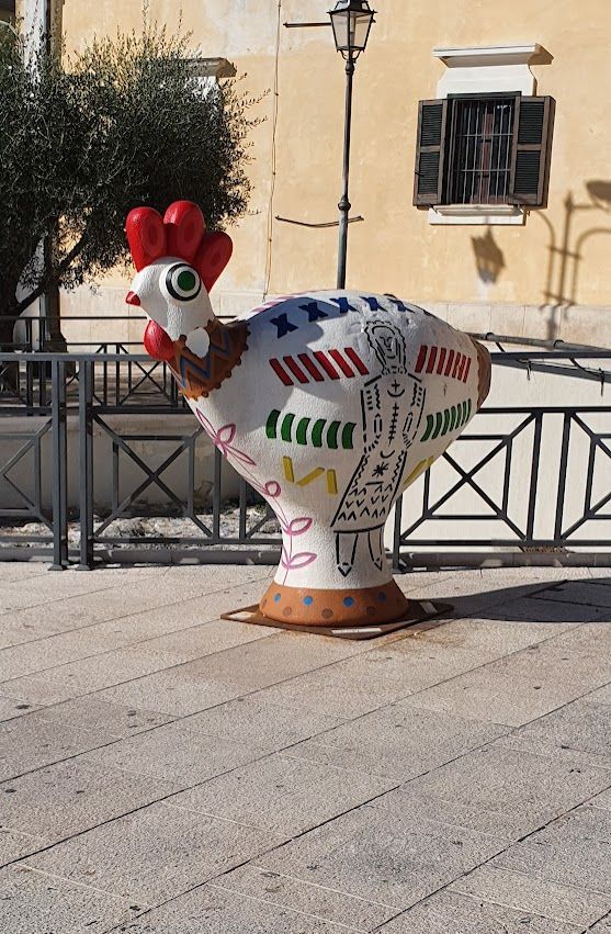 A colorful, artistic chicken sculpture with painted patterns, displayed outdoors against a historic European-style building.