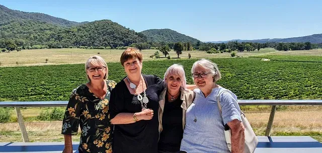 Four smiling women posing on a balcony overlooking a vineyard with rolling green hills.