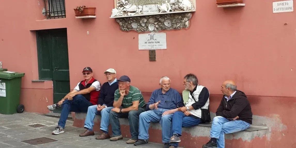 Men sitting and chatting on a bench by a red wall in a quiet Italian town.