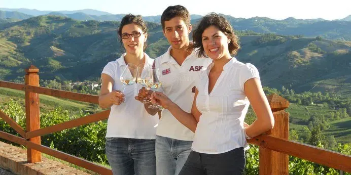 Three people holding wine glasses and smiling, with a scenic vineyard and rolling hills in the background.