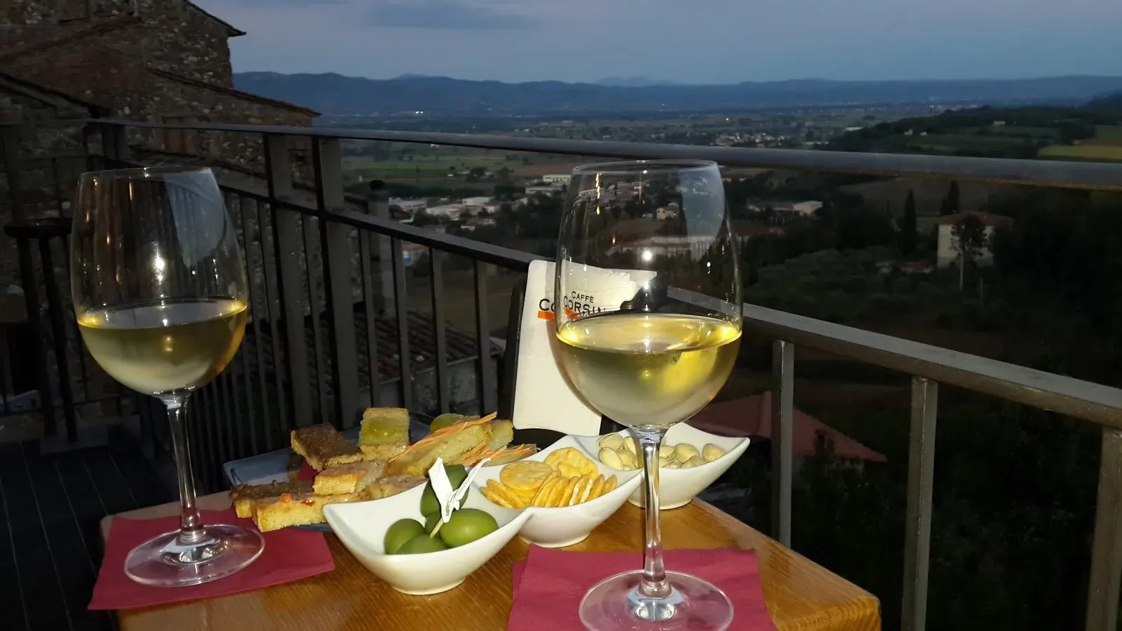 Two glasses of white wine with snacks on a balcony table overlooking a scenic valley at dusk.