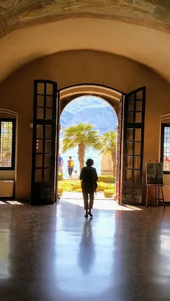 Silhouetted figure walking through arched doors of a historic building toward a sunlit courtyard.