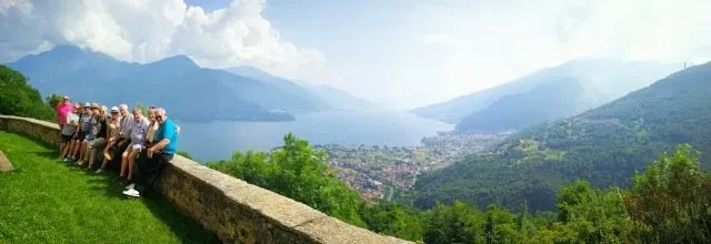 Tour group seated along a stone wall overlooking Lake Como, with mountains and lakeside towns visible under a hazy sky.