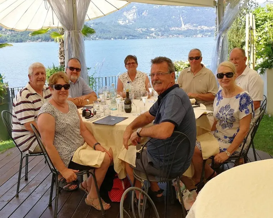 Eight people dining under a canopy with lake and mountain views in northern Italy. Table set with wine, glasses, and plates.