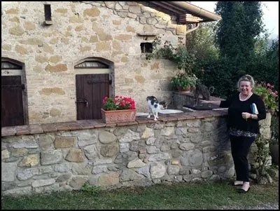 Woman standing beside a stone wall with a cat and potted flowers in front of a rustic brick building.