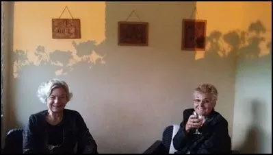 Two women sit indoors smiling, one holding a glass of wine, with wall shadows cast by plants in warm light.