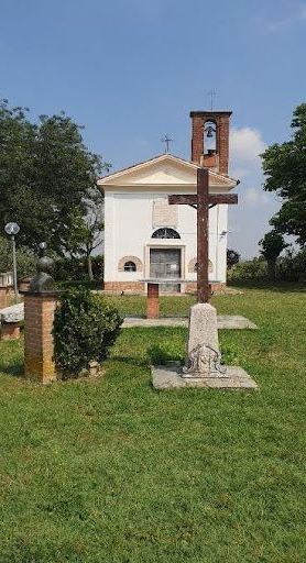 Small countryside church with a bell tower, wooden cross, and memorial stones on a grassy field.