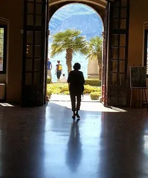 Silhouetted figure walking through arched doors toward a sunny lakeside garden with palm trees.