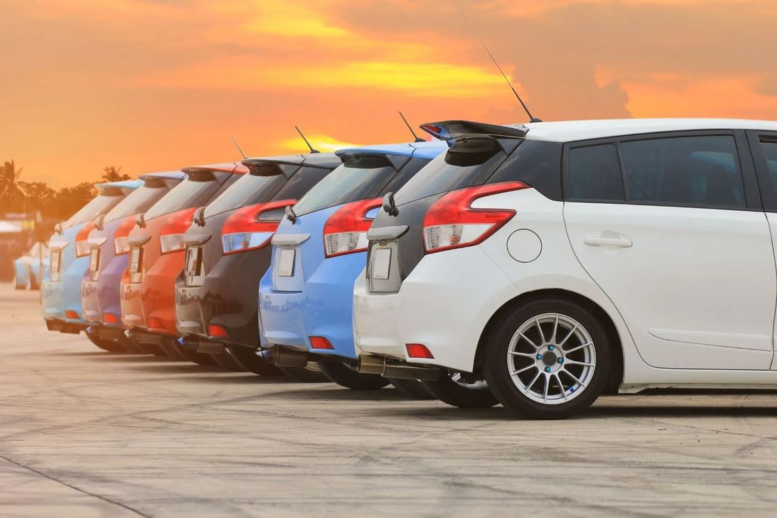 Una fila di auto hatchback parcheggiate, di colore arancione, grigio, blu e bianco, al tramonto.
