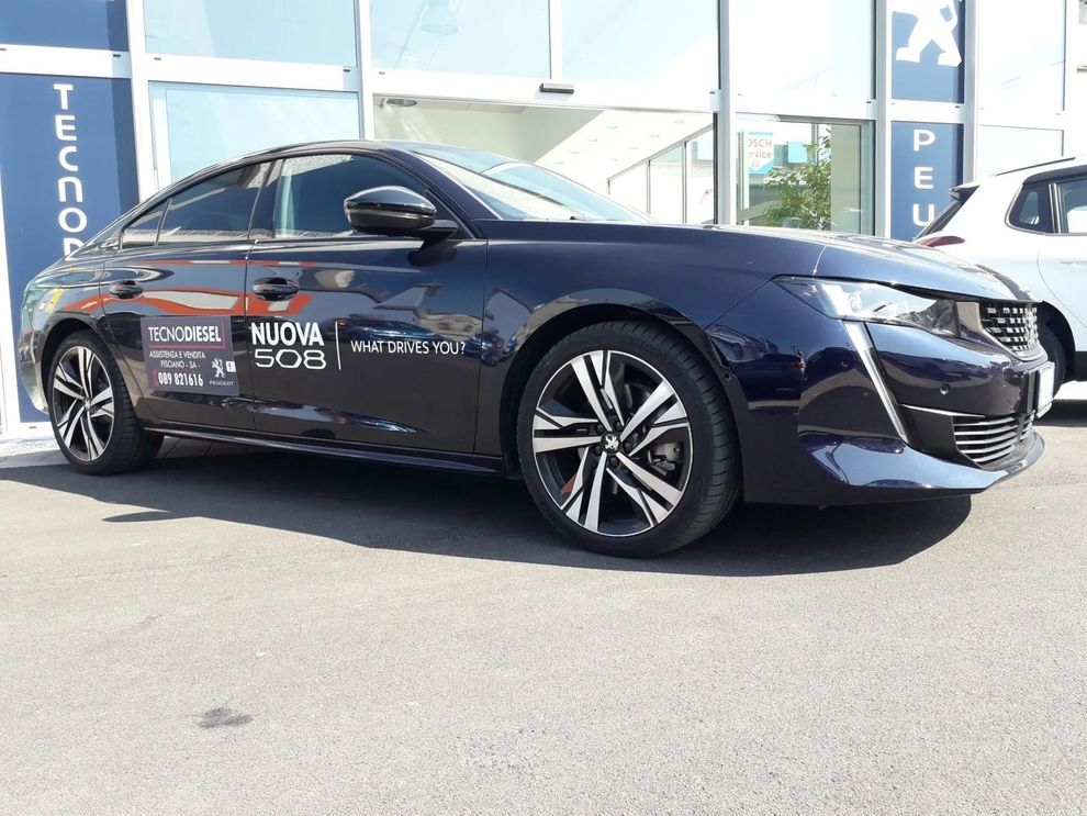 Berlina blu scuro parcheggiata fuori da una concessionaria con adesivi promozionali sulla fiancata.