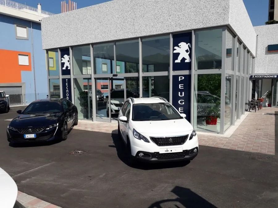 Due auto parcheggiate fuori da una concessionaria Peugeot con vetrina e insegna con logo.