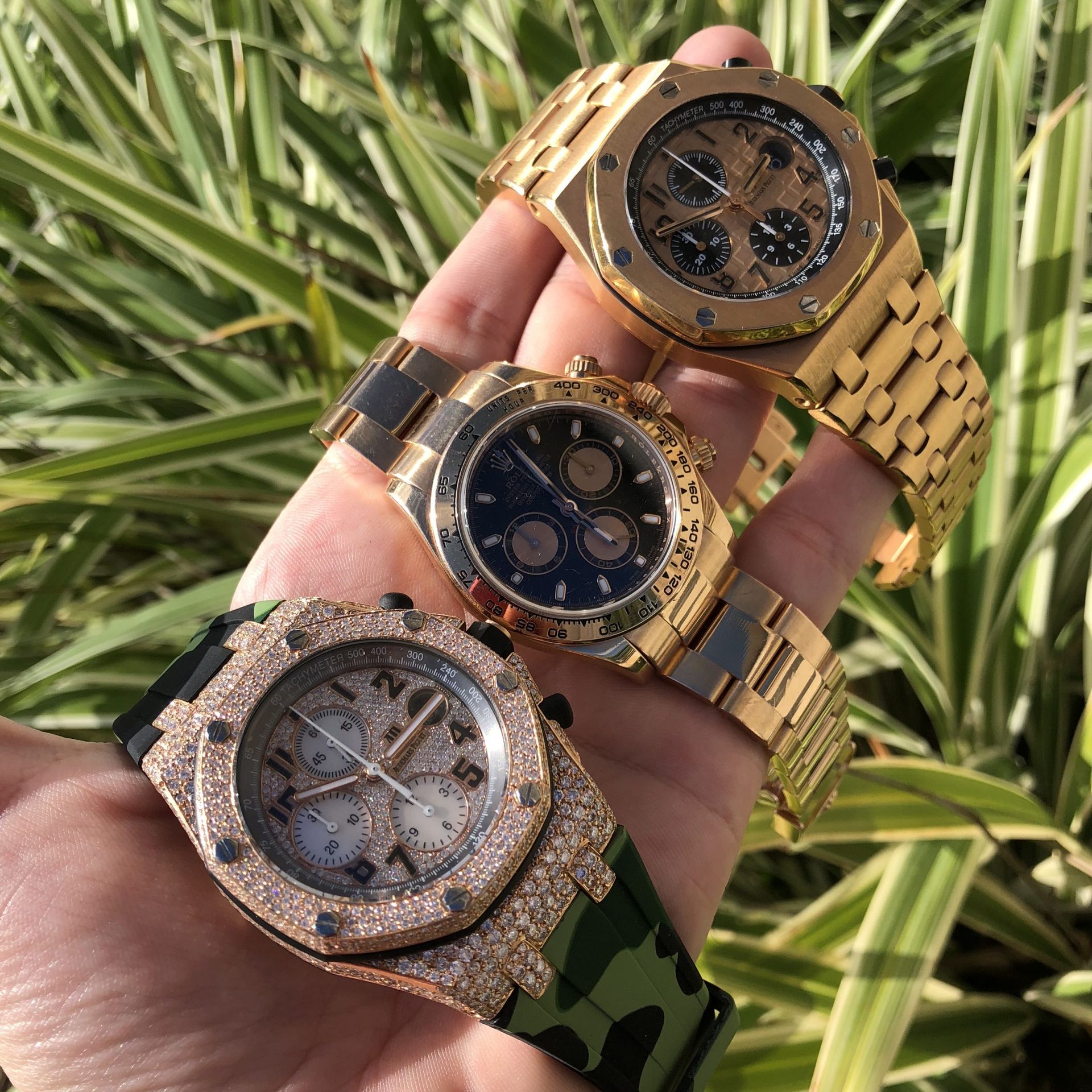 Person's hand holding three luxury gold watches: Audemars Piguet, Rolex Daytona, and camouflage strap watch with diamond bezel.