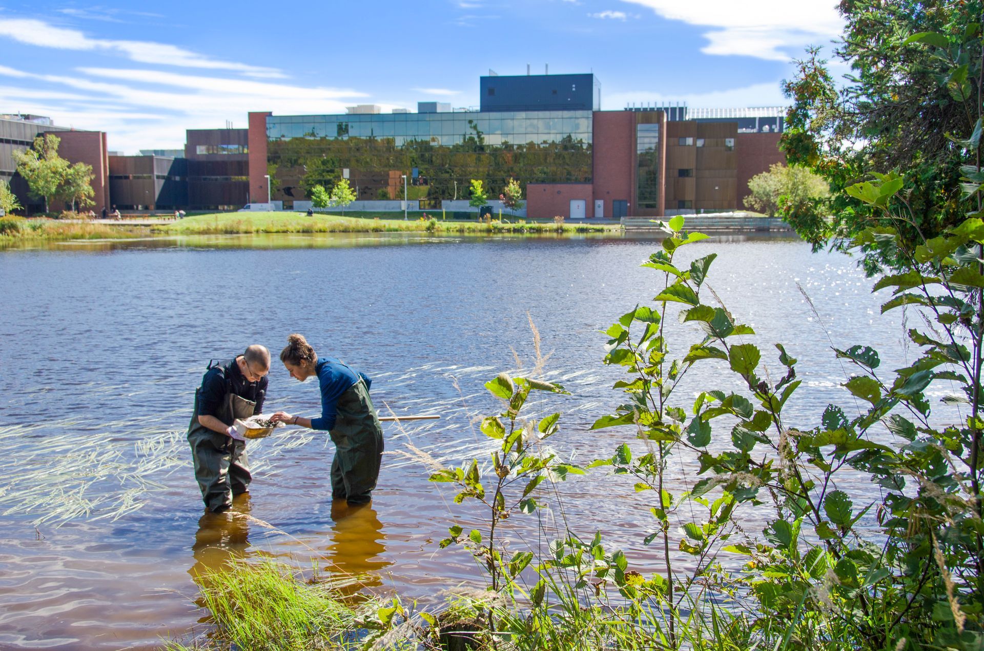 Two people in waders collect water samples from a lake; Nipissing University building in the background.