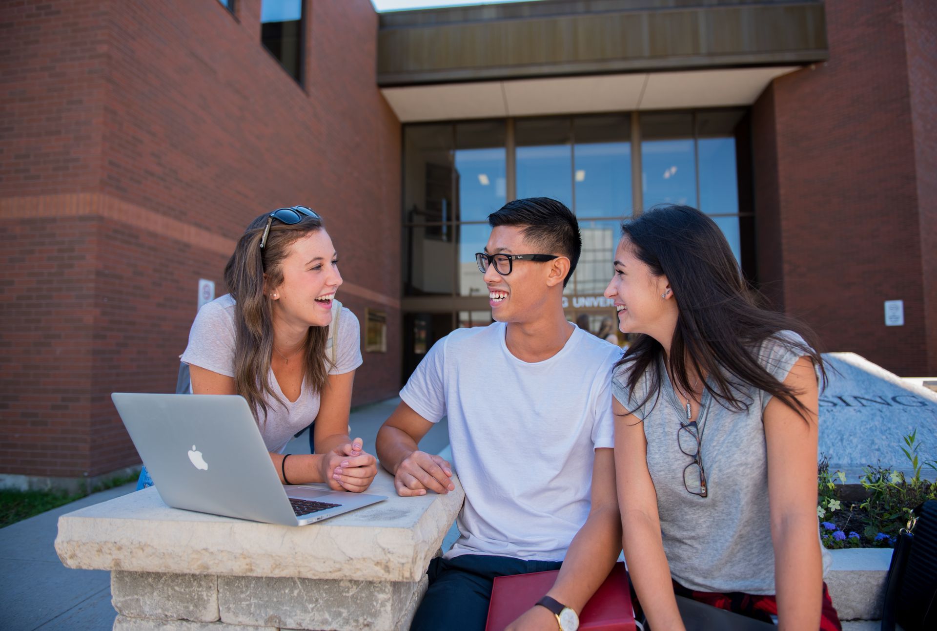 Three students outside Nipissing University building, looking at laptop, smiling and talking.