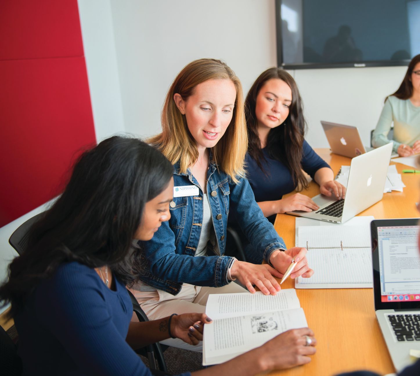 Group of women reviewing papers around a table, laptop computers, in a modern office with a red wall.