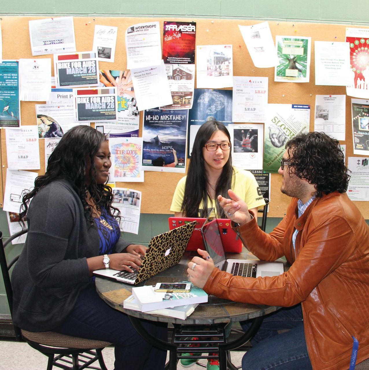 Three people around a table, looking at a laptop and discussing in front of a bulletin board.
