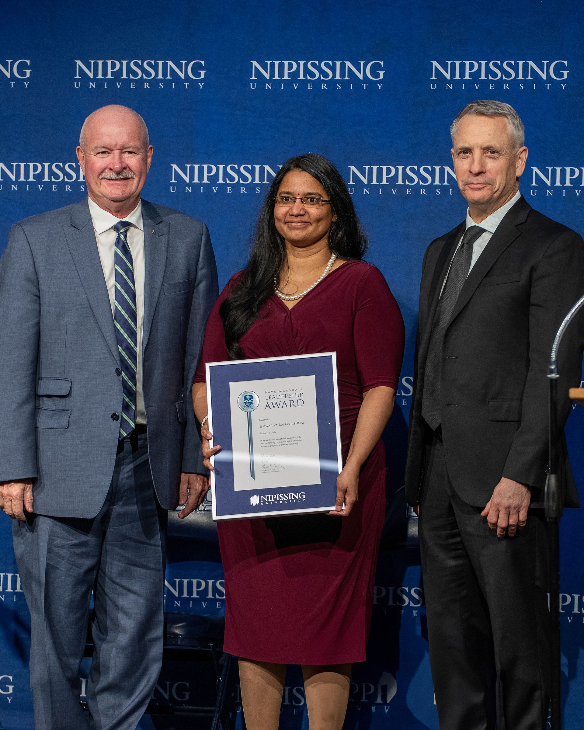 Three people at Nipissing University event. A woman in a red dress holds a framed award.