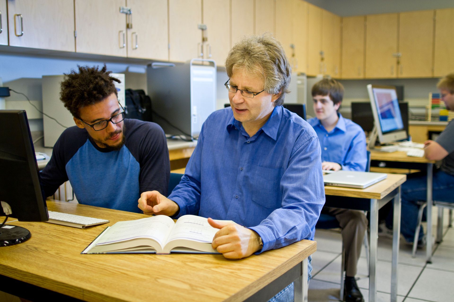 Professor assisting a student at a computer, other students work in a classroom setting.