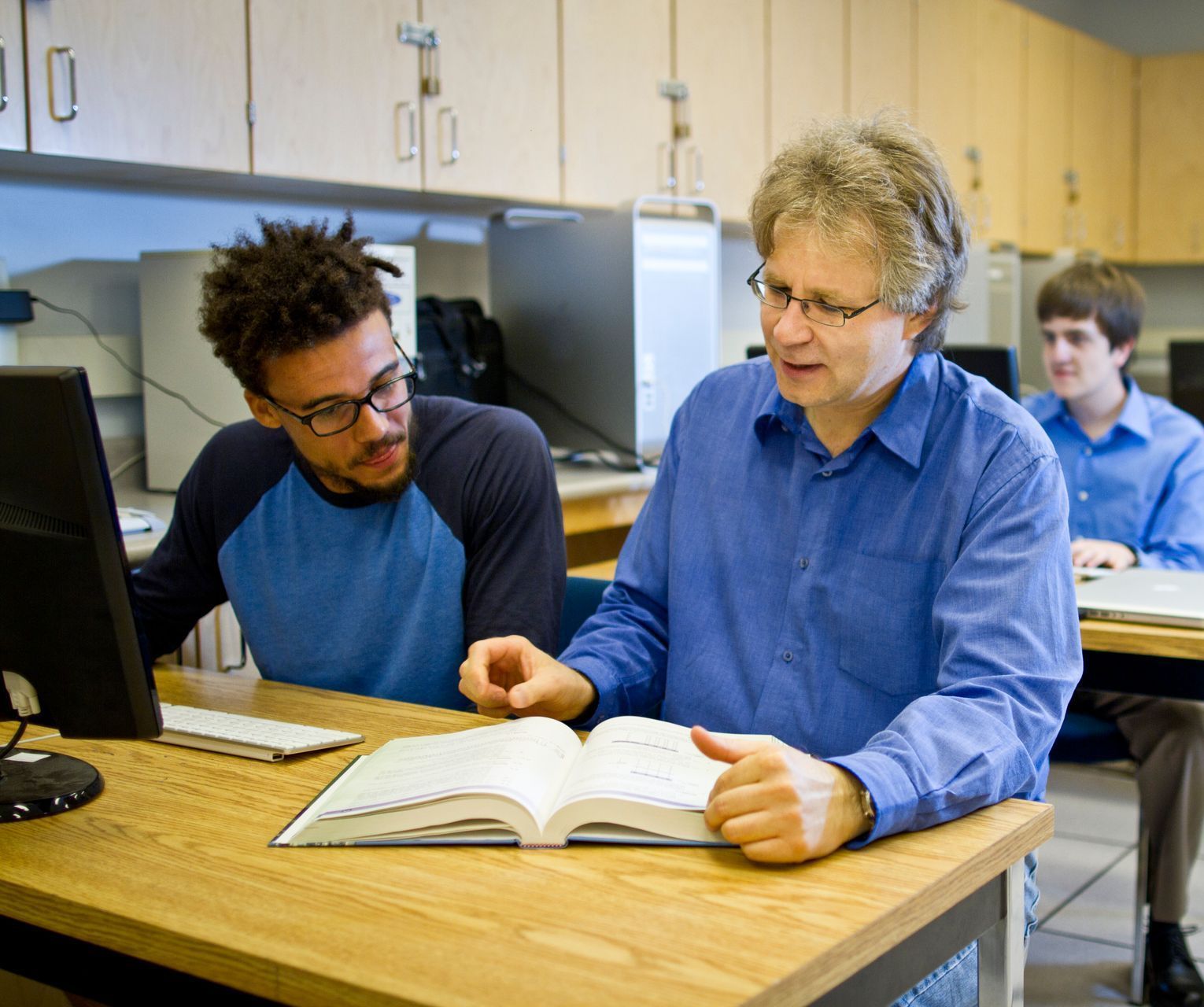 A professor and a student look at a book together in a computer lab. Another student works in the background.