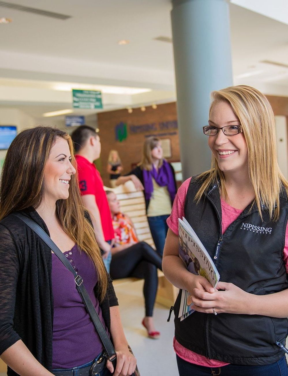 Two smiling young women talking inside a building. Others are in the background.