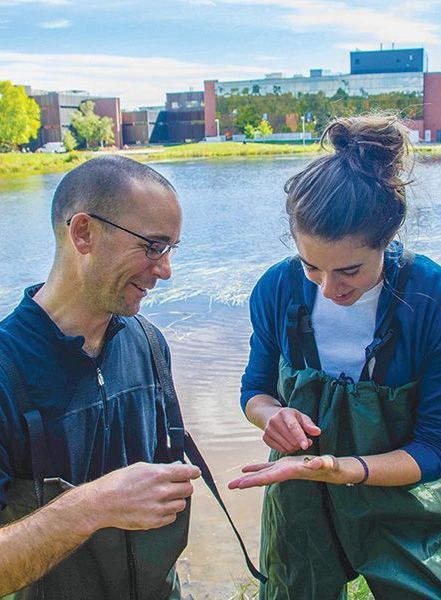 A man and woman in waders by a lake, examining an item in their hands; campus buildings in background.