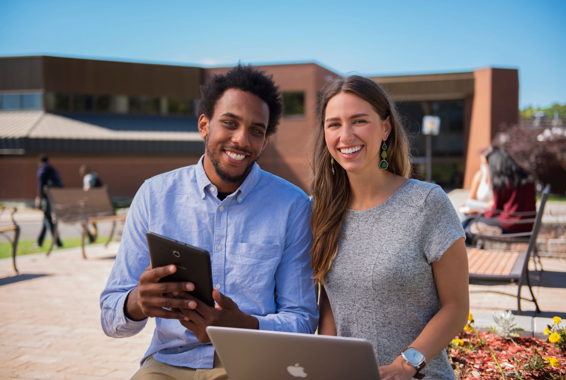 Two students smiling outdoors with a laptop and tablet, in front of a brick building.