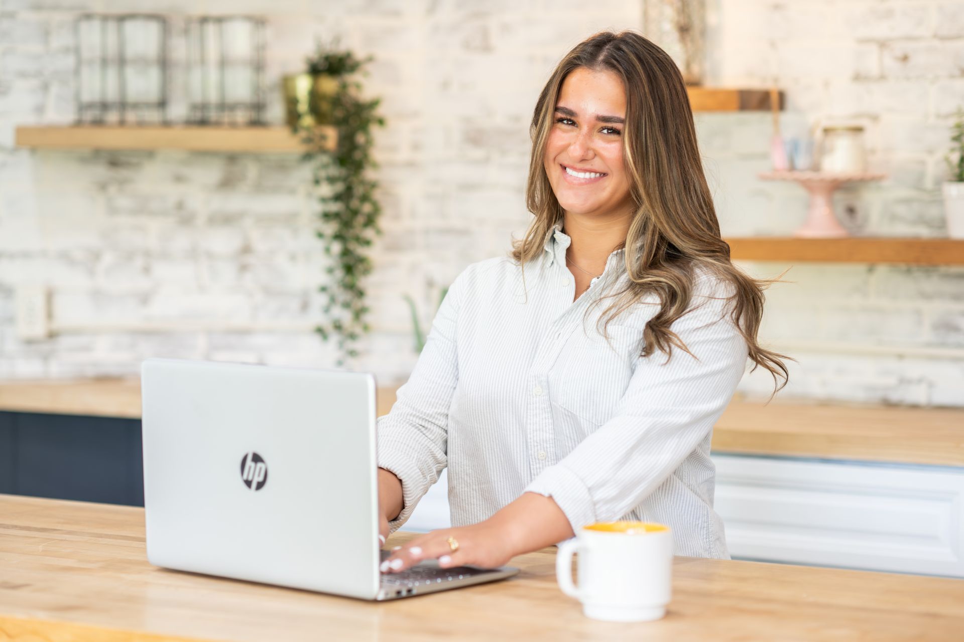 Woman smiling, typing on a laptop, in a kitchen setting with a mug nearby.