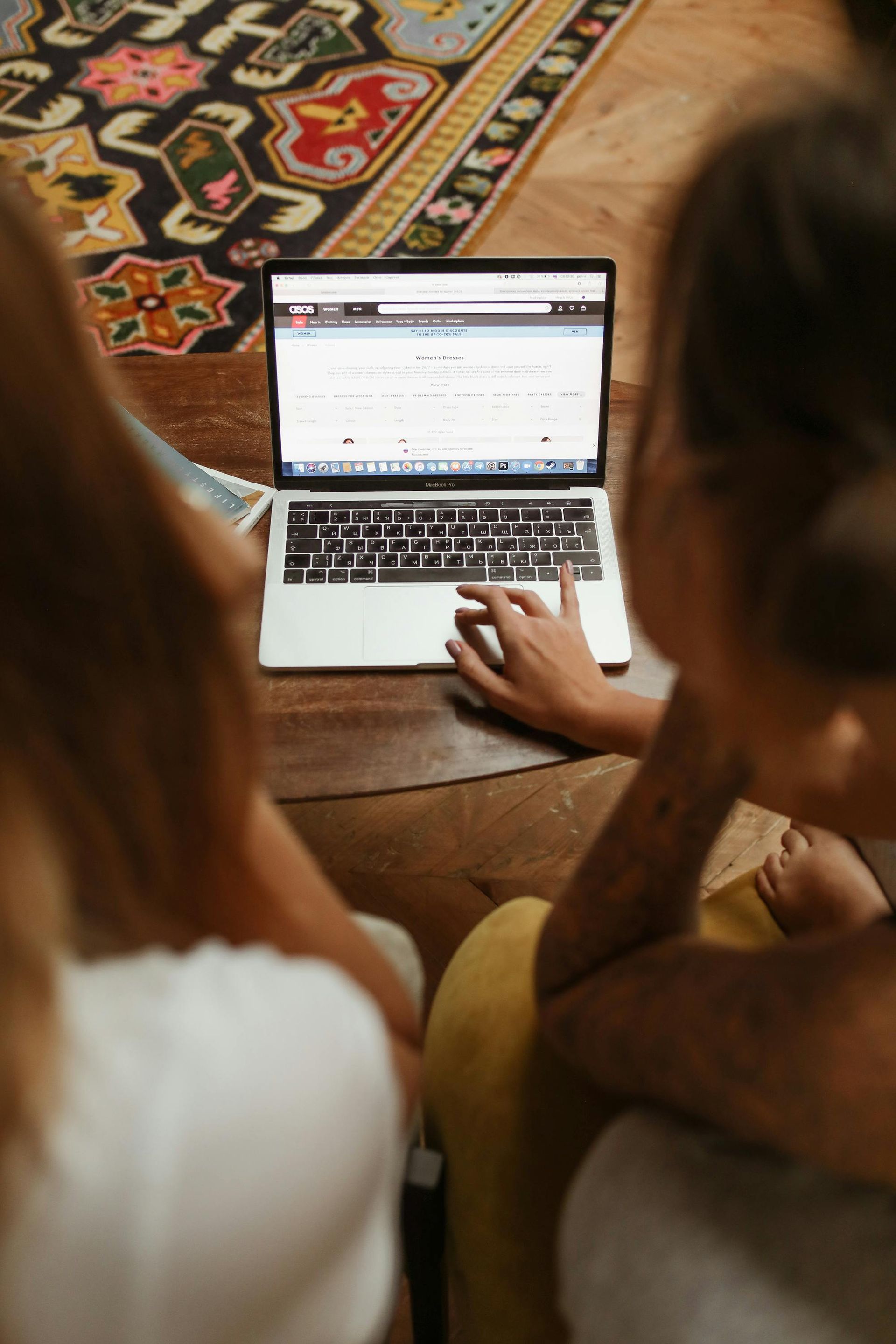 Two people looking at a laptop on a wooden floor with a colorful rug. One person types.